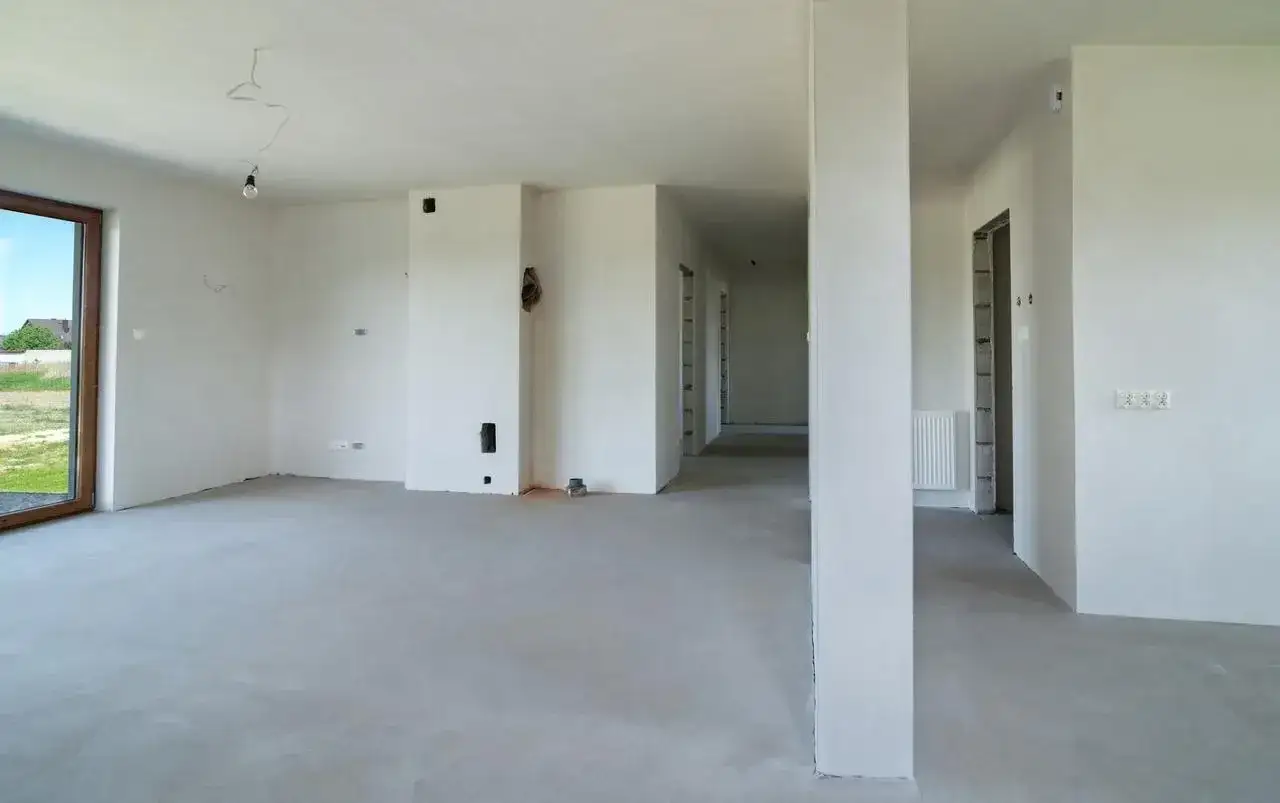Wnętrze domu w stanie deweloperskim: białe ściany, wylewka na podłodze, widoczne instalacje, drzwi do pomieszczeń.