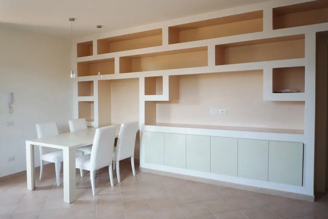 Un moderno soggiorno con tavolo bianco e sedie, dominato da un originale muretto in cartongesso con vani a giorno e pensili a scomparsa.