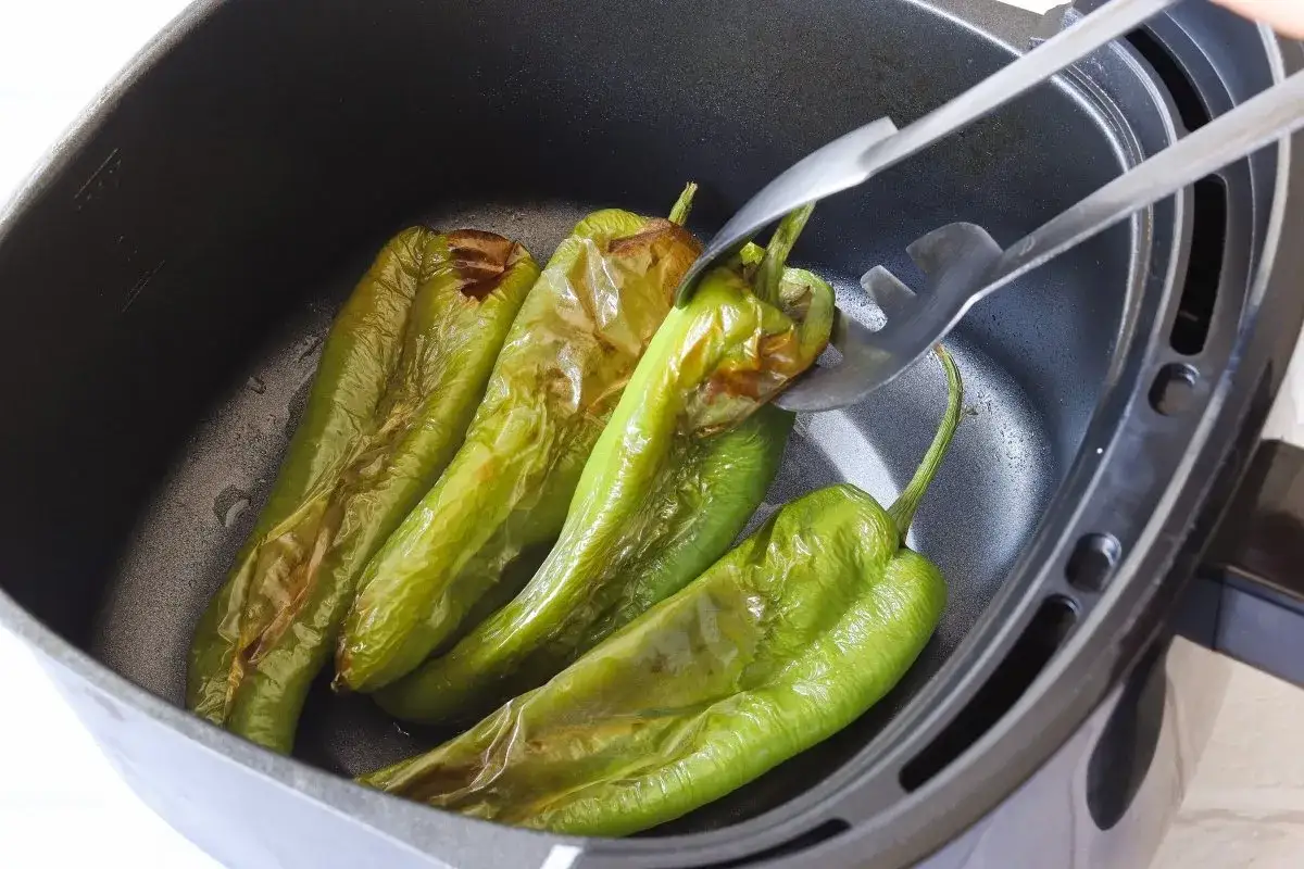 Pimientos verdes asándose en la freidora de aire. Unas pinzas voltean uno de los pimientos para que se cocine uniformemente.