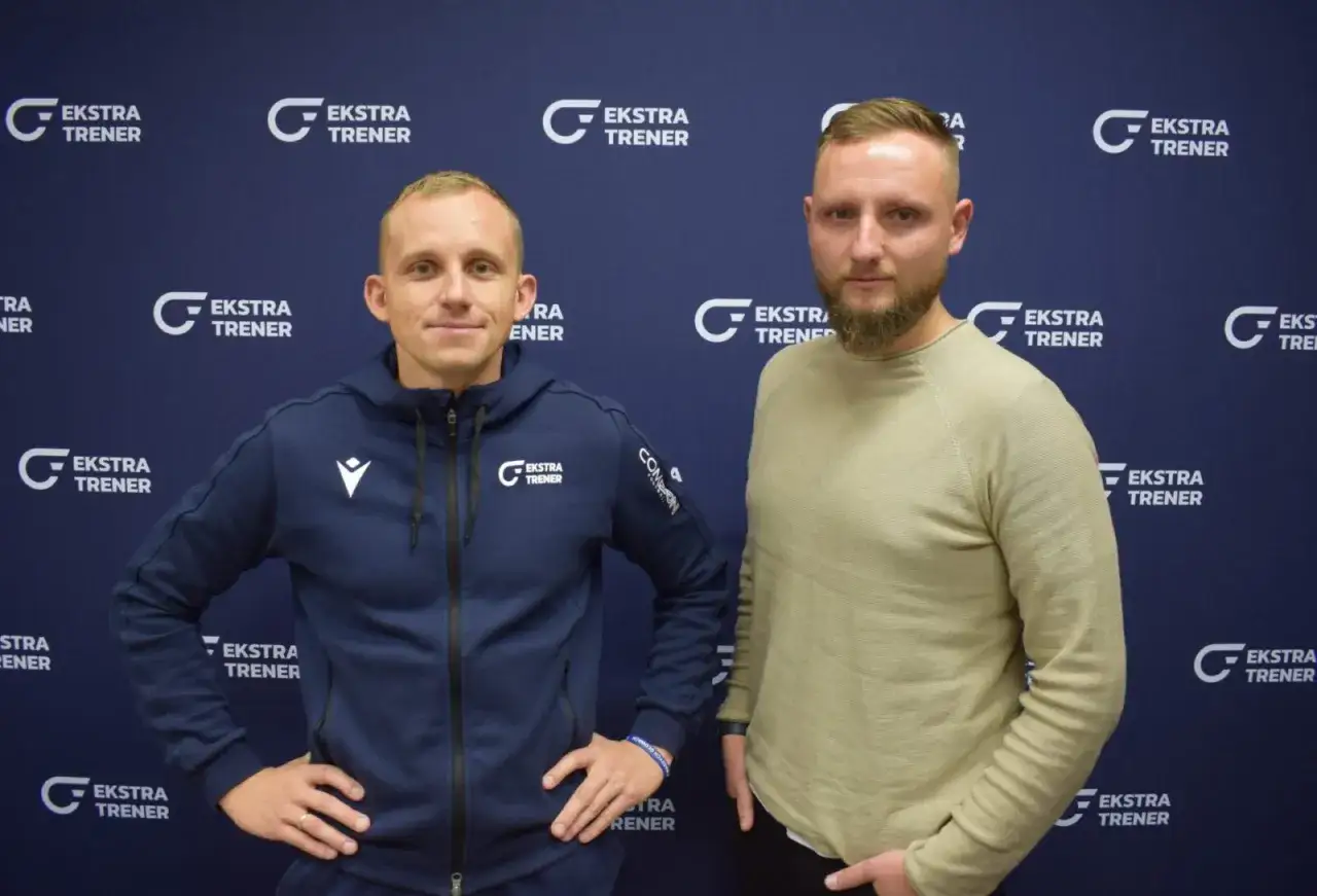 Dwóch mężczyzn, jeden w granatowej bluzie z kapturem, drugi w beżowym swetrze, pozuje na tle z logo