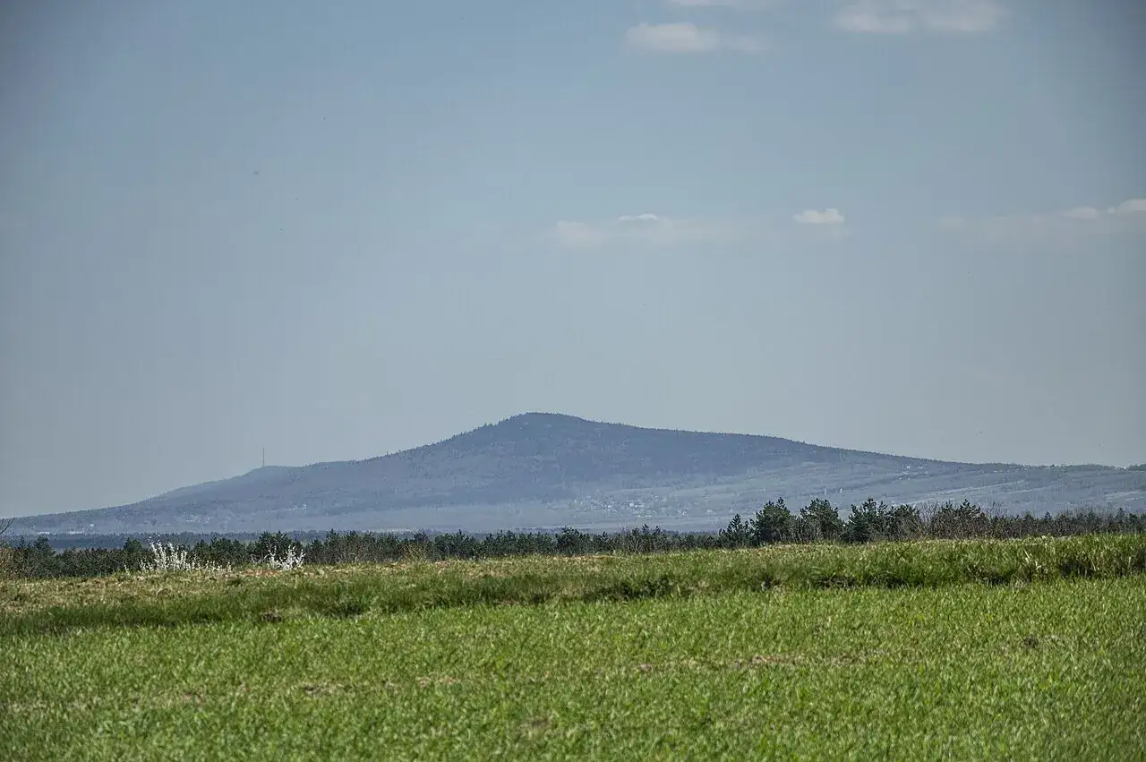 Łagodne, zalesione góry świętokrzyskie szczyty w oddali, na tle błękitnego nieba z chmurkami. Na pierwszym planie zielona łąka.