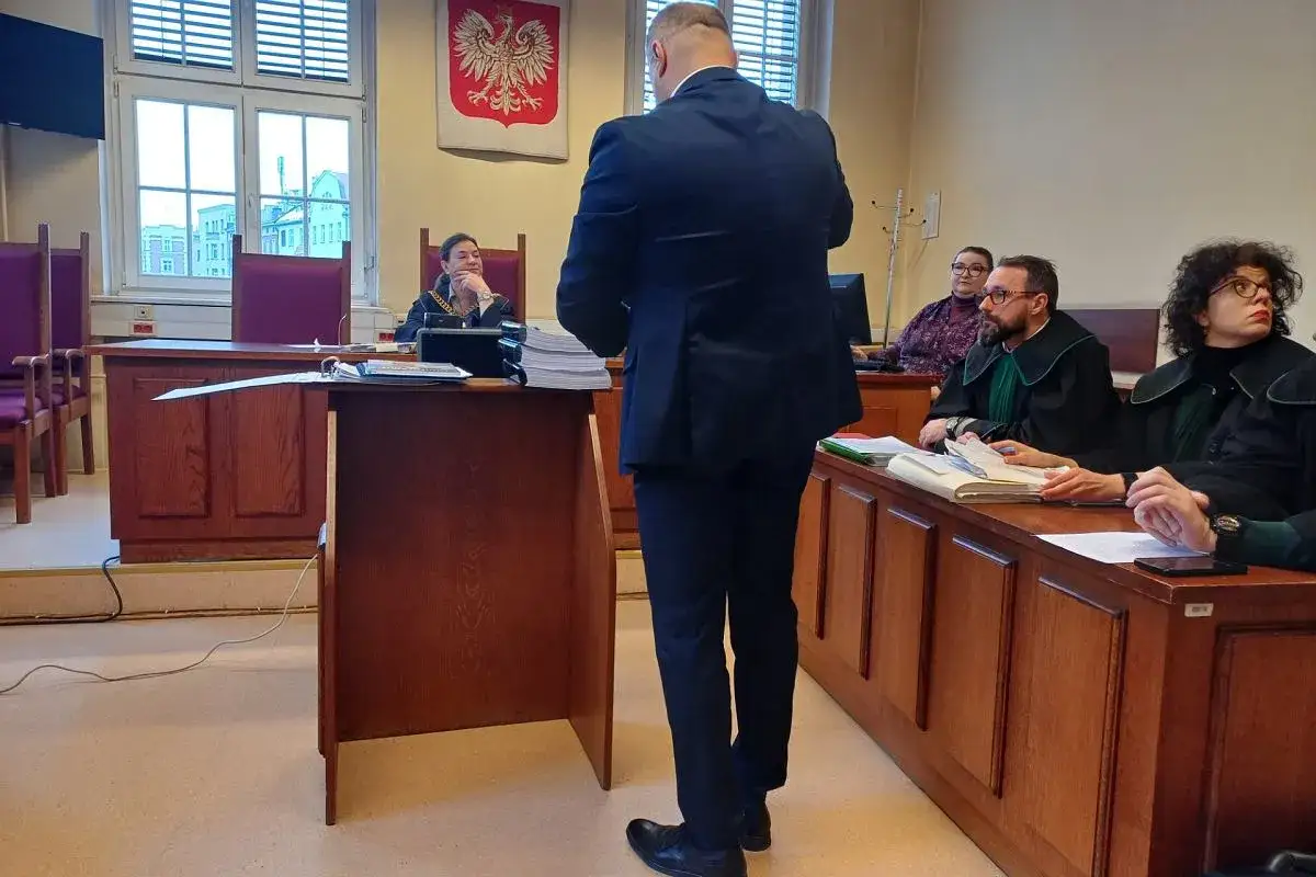 Mężczyzna w garniturze stoi przed ławą sędziowską, gdzie siedzi sędzia i prokurator. Rozważane jest dobrowolne poddanie się karze prokurator.