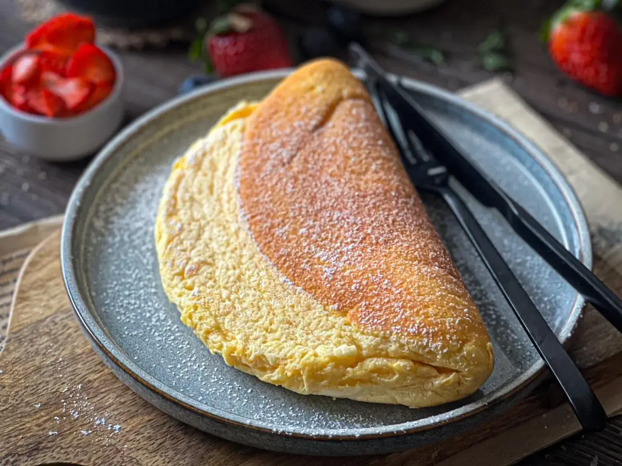 Puszysty omlet na talerzu, posypany cukrem pudrem. Idealny przykład, jak zrobić omlet, który zachwyci smakiem i wyglądem. Obok widoczne truskawki.