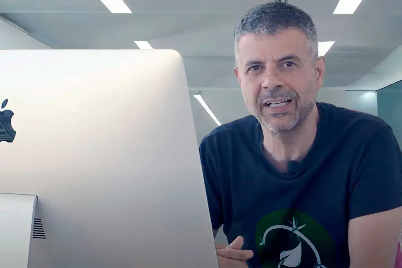 Hombre con barba gris habla frente a un iMac. Podr&iacute;a ser un asesor energ&eacute;tico explicando c&oacute;mo optimizar el consumo.