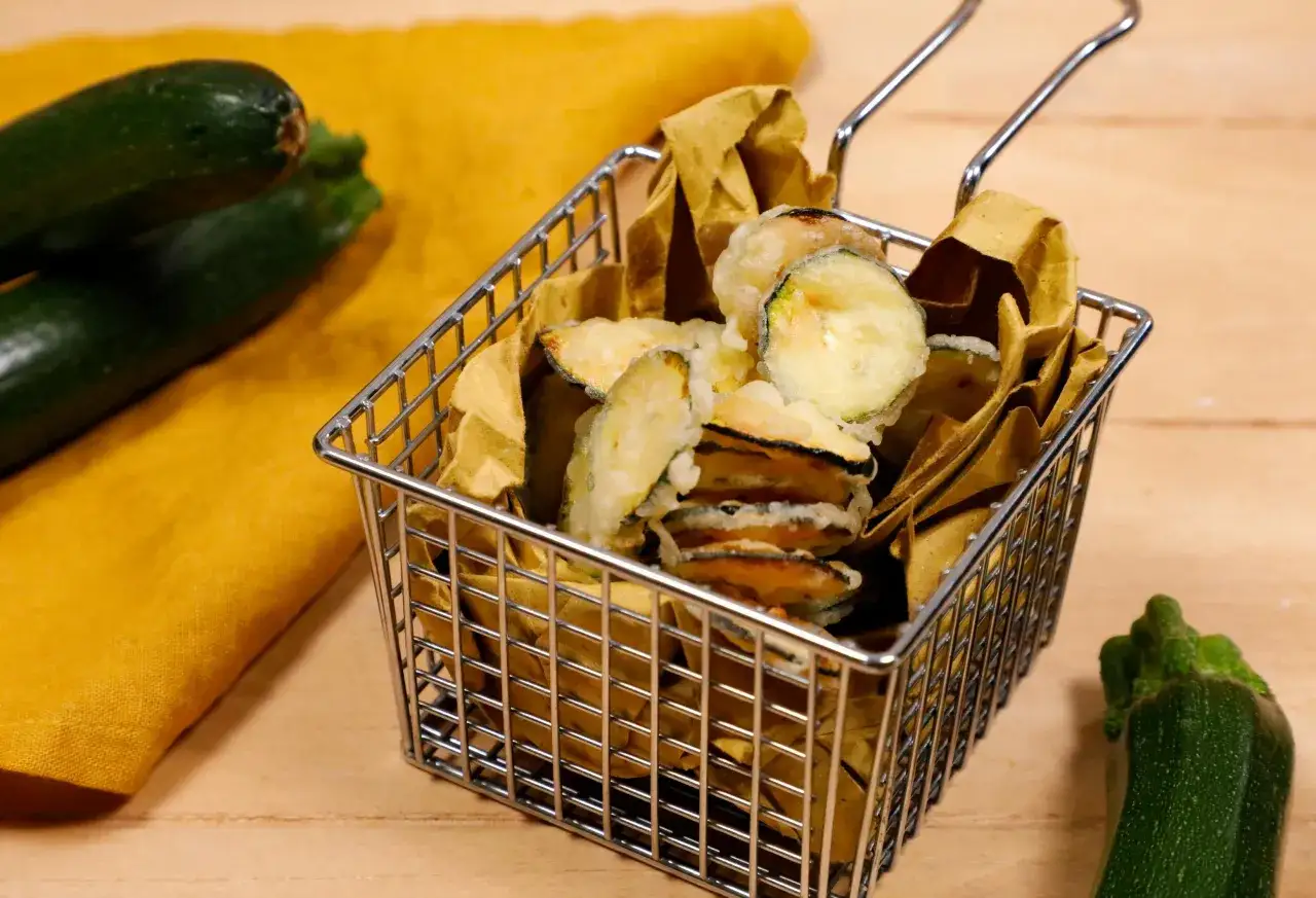 Fettine dorate di zucchine in pastella, servite in un cestino metallico, con zucchine fresche e un tovagliolo giallo sullo sfondo.