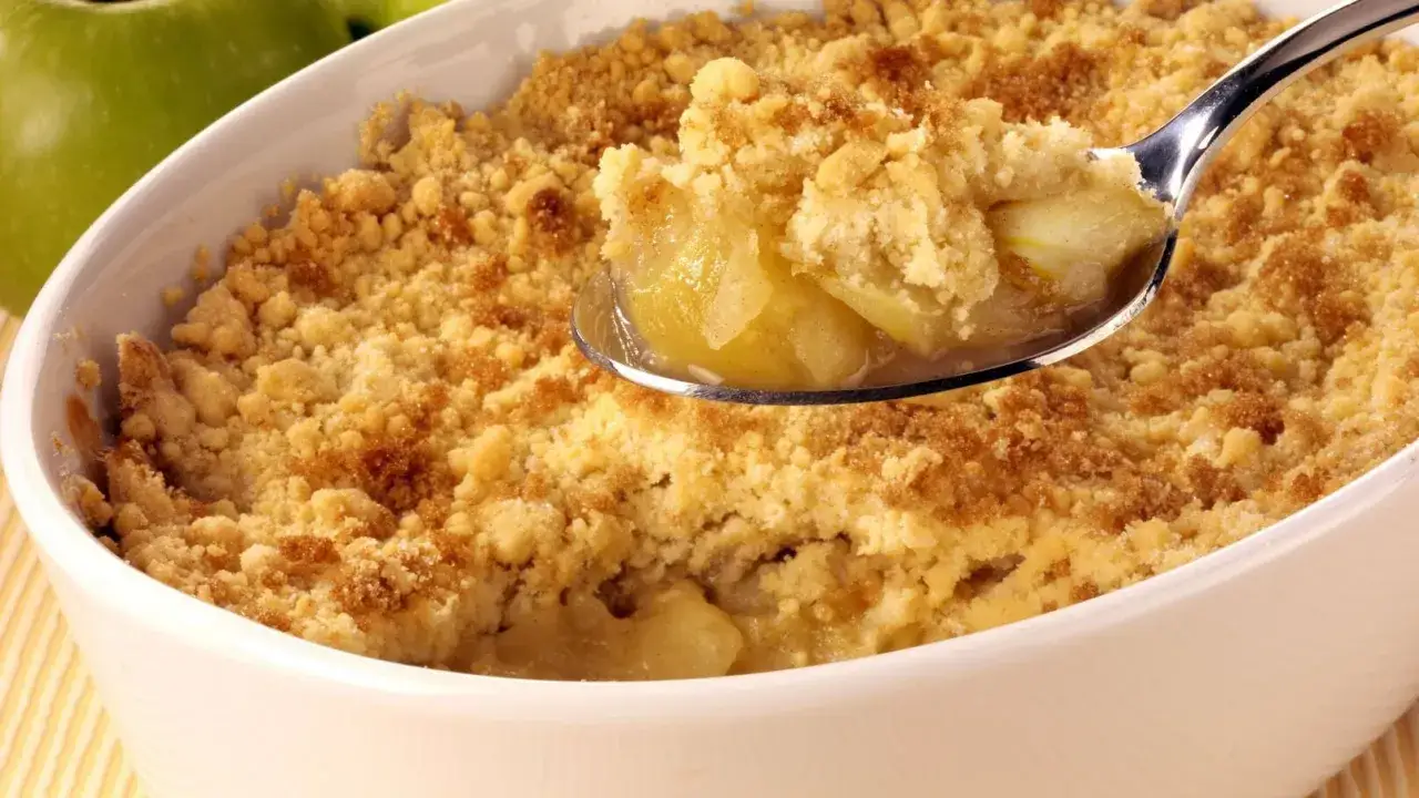 Un délicieux crumble aux pommes, parfait pour suivre la recette de Cyril Lignac. Une cuillère en argent prélève une bouchée gourmande.