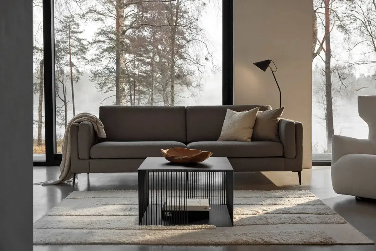 En stilren soffgrupp med en gr&aring; soffa och en svart soffbordskub, placerad framf&ouml;r ett stort f&ouml;nster med utsikt &ouml;ver en skogssj&ouml;.