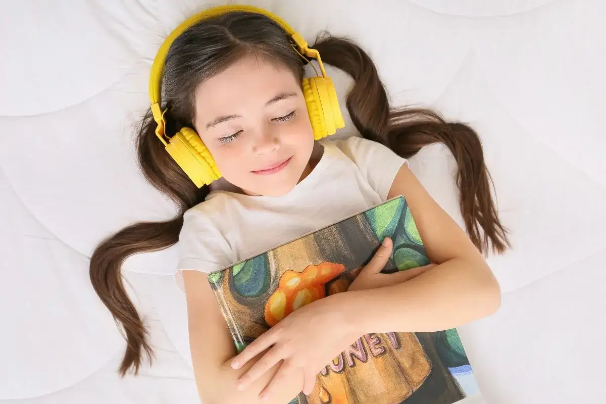 Dziewczynka w ż&oacute;łtych słuchawkach leży z zamkniętymi oczami, przytulając książkę. Słucha audiobook&oacute;w, zanurzając się w świat opowieści.
