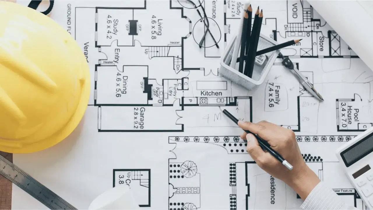 Un arquitecto o proyectista de una obra revisa planos con un casco amarillo, lápices y calculadora.