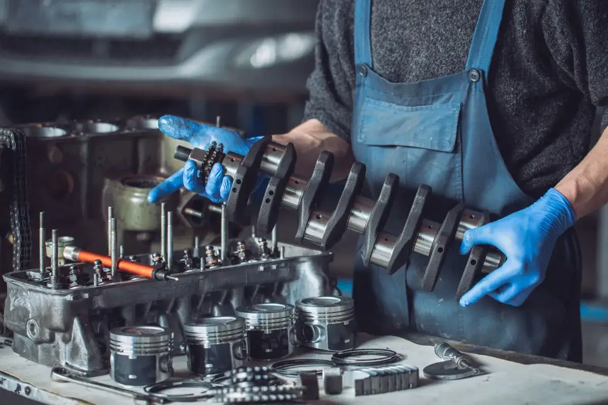 Mechanik w niebieskim kombinezonie trzyma wał korbowy silnika 1.4 tdi. Na stole leżą tłoki i inne części silnika.