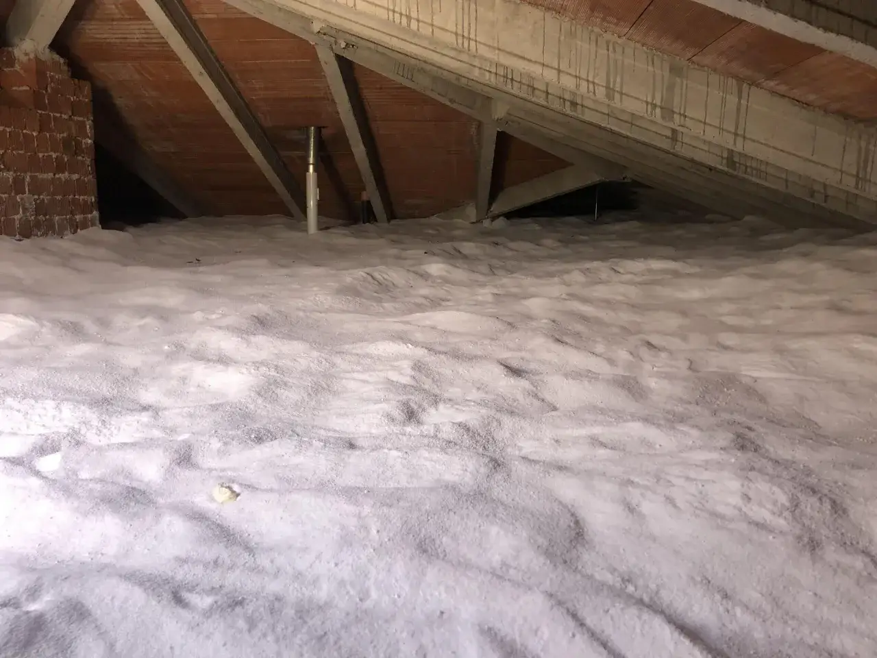 Sottotetto con isolamento a fiocchi bianchi, simile a neve, che copre il pavimento. Travi in legno e cemento visibili sopra.
