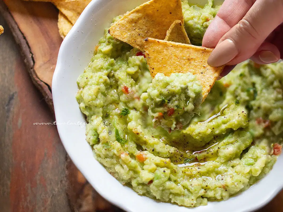 Una mano intinge un chip di mais in una ciotola di guacamole fresco, mostrando come fare il guacamole fatto in casa.