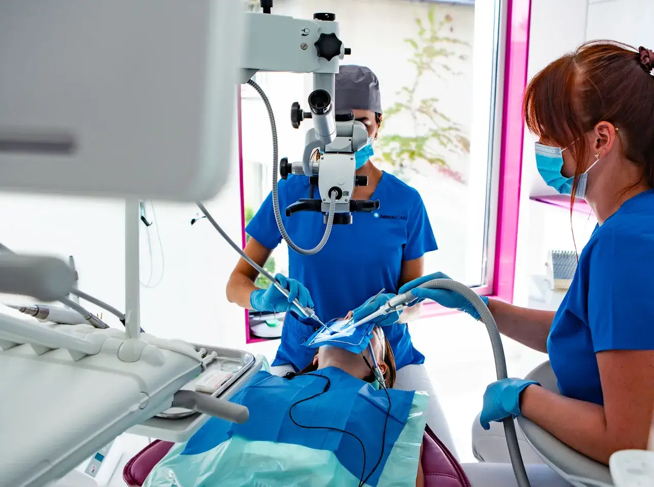 Dentystka pod mikroskopem leczy pacjentowi ząb, kt&oacute;ry m&oacute;głby być siny. Asystentka podaje narzędzia.
