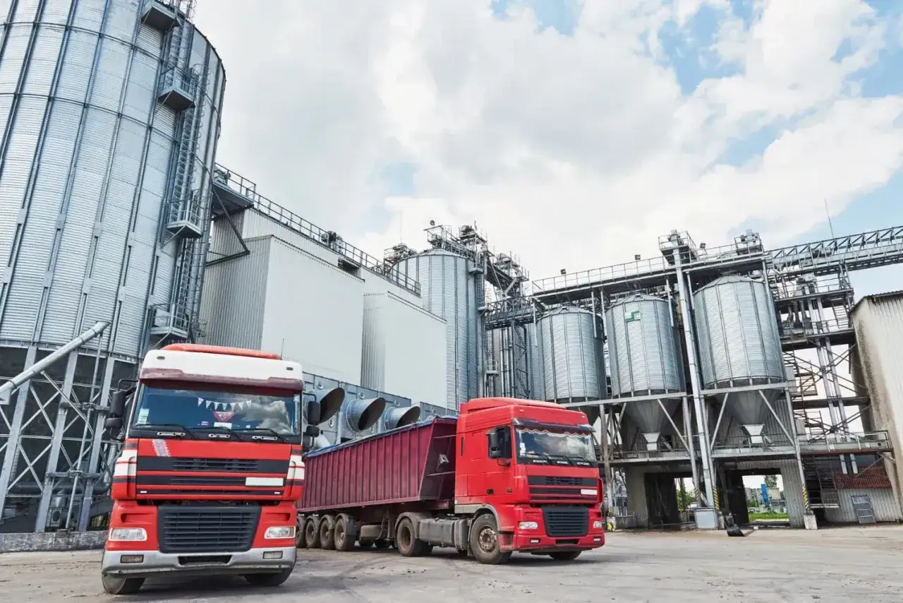 Dwa czerwone ciężarówki z naczepami stoją przed silosami zbożowymi. Transport ponadgabarytowy, godziny jazdy.