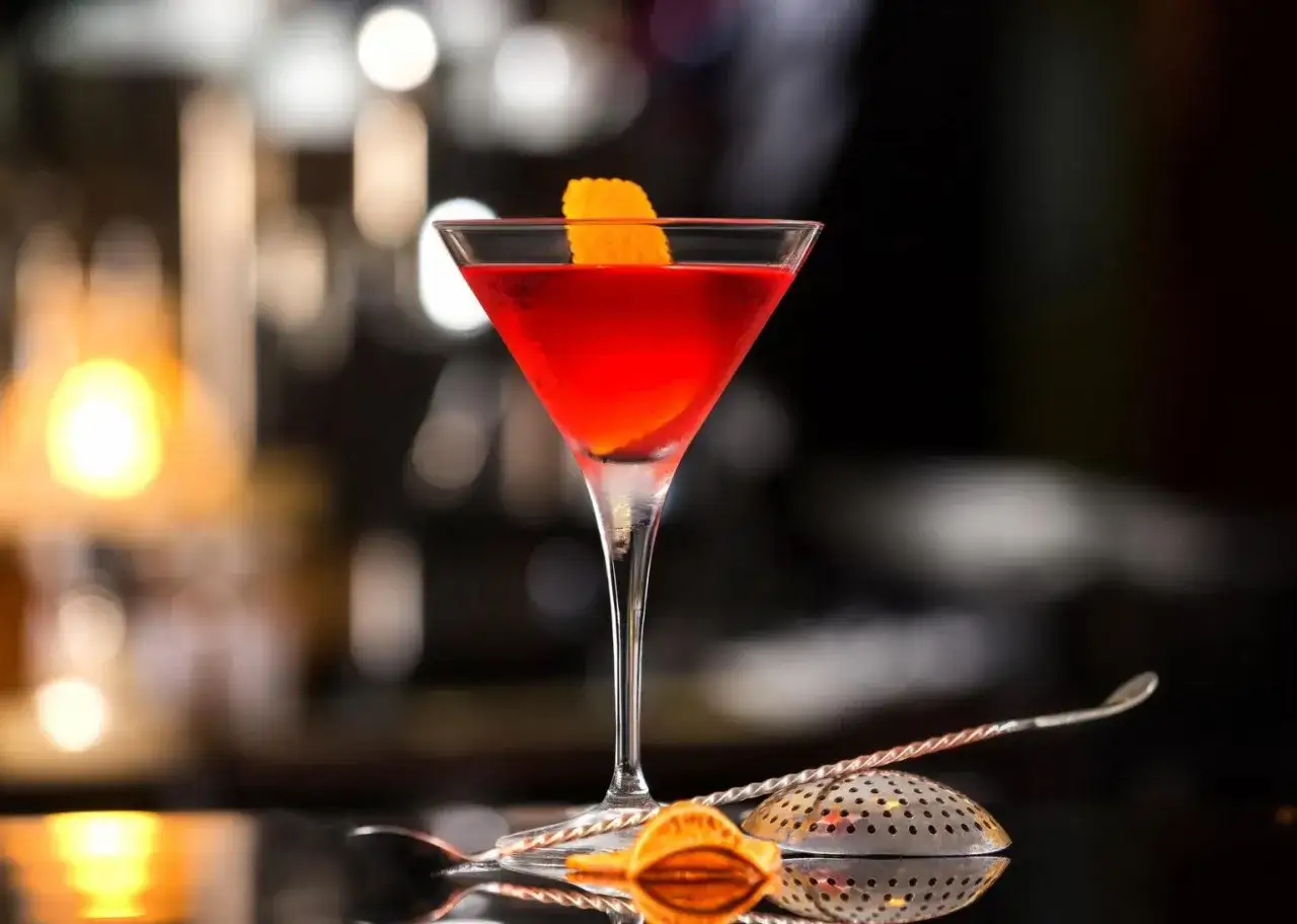Czerwony Manhattan drink w kieliszku, ozdobiony skórką pomarańczy. Obok leży barmańska łyżka i sitko.
