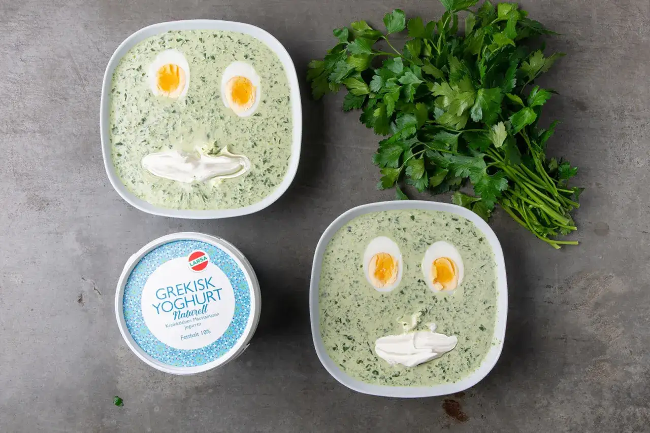 Två skålar med spenatsoppa dekorerade som ansikten, med kokta ägg som ögon och gräddfil som mun. Bredvid en kruka grekisk yoghurt och en bukett persilja. Ett roligt spenatsoppa recept för barn.