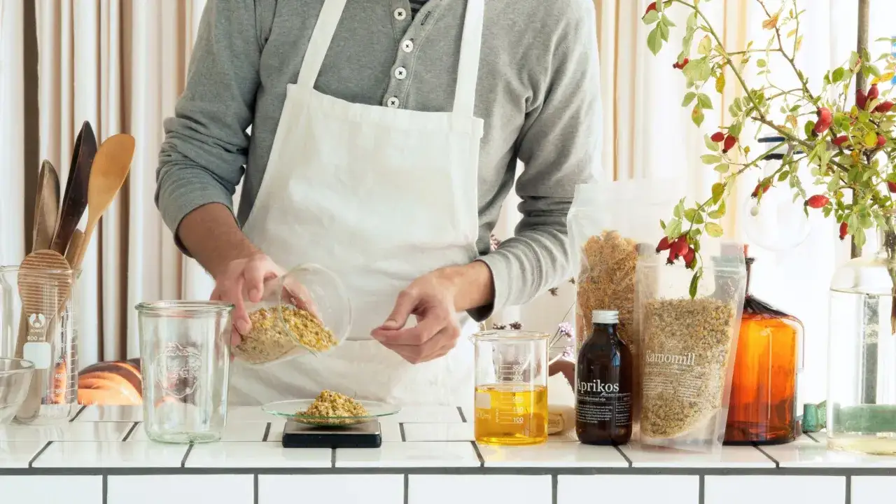Person i f&ouml;rkl&auml;de m&auml;ter upp torkad kamomill f&ouml;r att g&ouml;ra vegetabilisk olja. Bredvid st&aring;r en flaska aprikosk&auml;rnolja och andra ingredienser.