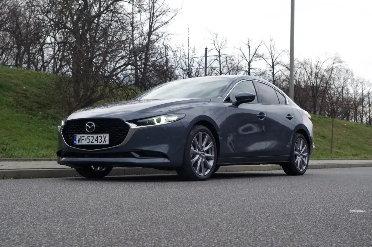 Szary sedan Mazda 3 2.0 150KM na tle drzew. Wiele osób szuka takich aut, bo mazda 3 2.0 150km opinie ma dobre.