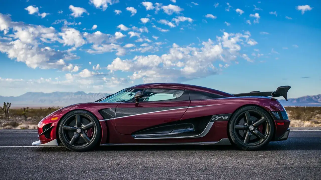 Ciemnoczerwony Koenigsegg Agera RS z karbonowymi detalami i masywnym skrzydłem, zaparkowany na pustynnej drodze pod błękitnym niebem.