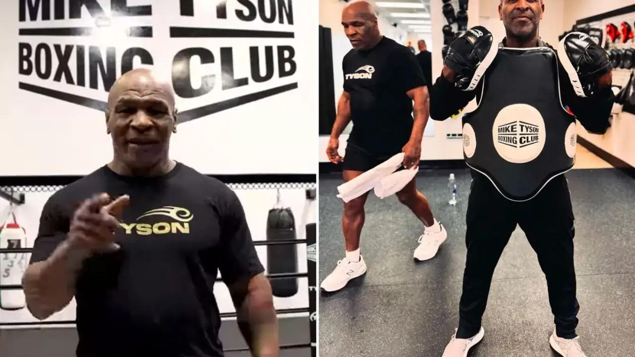 Mike Tyson in a boxing gym, possibly outlining his training schedule. He's wearing a black t-shirt with his logo. Another man is in the background wearing a chest protector.