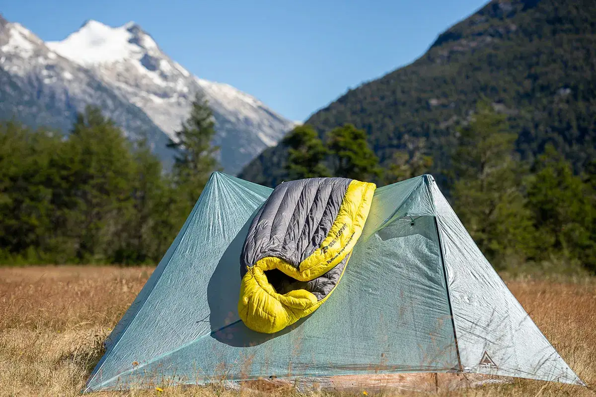 A gray and yellow Nemo Disco 15 sleeping bag rests on a light blue tent, set against a backdrop of forested mountains and a snowy peak.