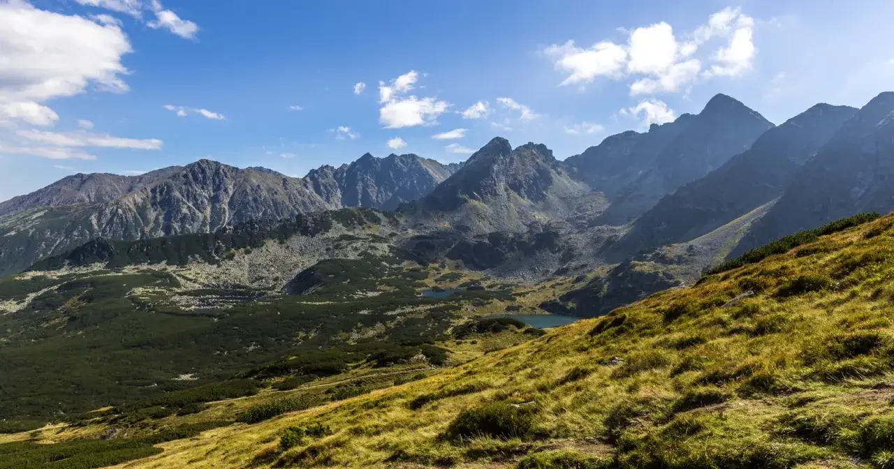 Widok na najwyższe góry w Polsce, Tatry, z trawiastego zbocza. W oddali widać skaliste szczyty i jeziora.