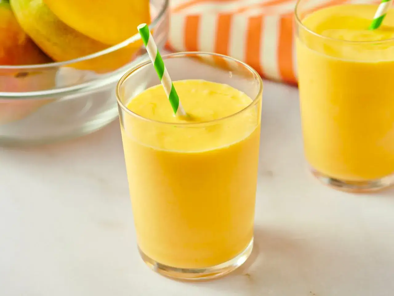 Orzeźwiający koktajl z mango w szklance z zielono-białą słomką. W tle miska z owocami mango.