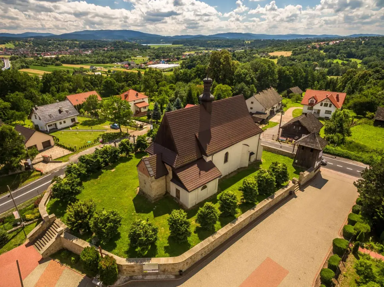 Widok z lotu ptaka na kościół i zabudowania w Dobczycach. Rynek otoczony zielenią, w tle góry.