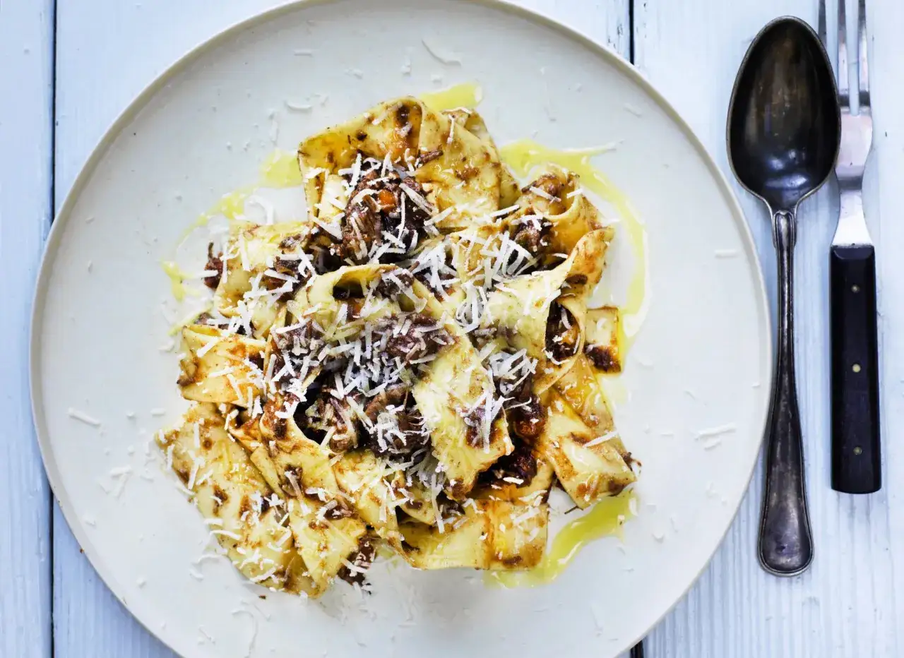 En tallrik med pappardelle al cinghiale, breda band av pasta med en mustig viltsås och riven parmesan. En sked och gaffel ligger bredvid.
