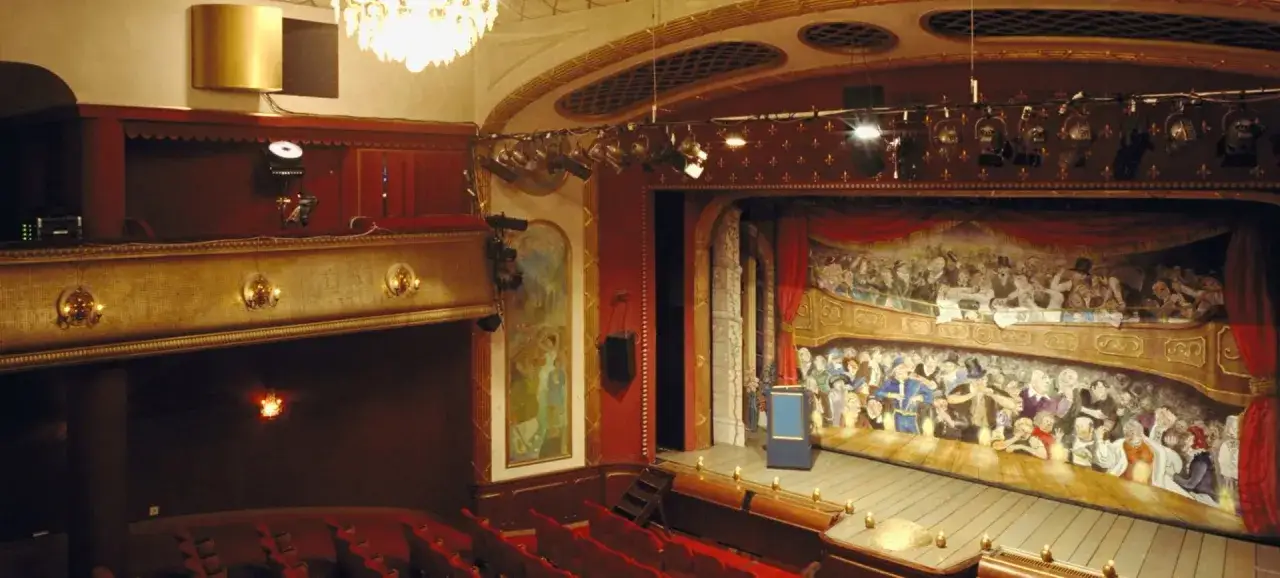 Teater Odenplan med sin pampiga scenm&aring;lning av publik i loger. R&ouml;da sammetssoffor och gyllene detaljer skapar en klassisk atmosf&auml;r.