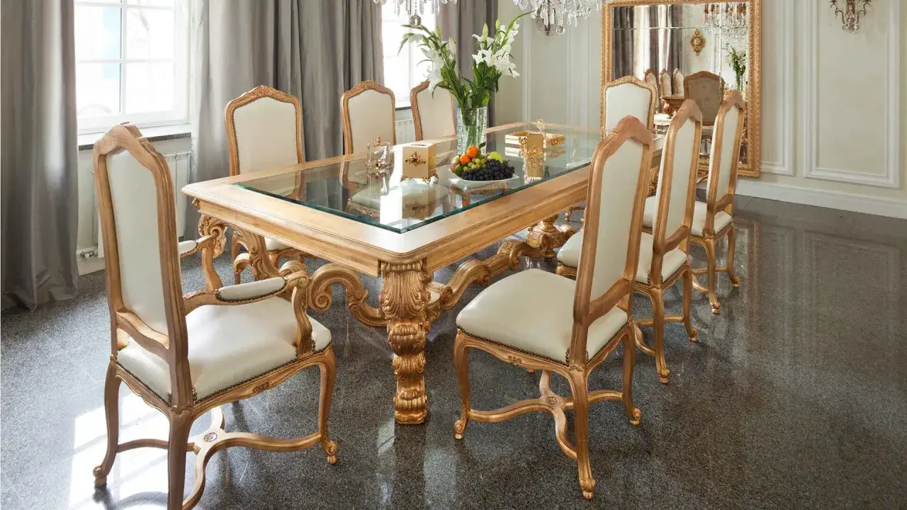 Tavolo da pranzo in legno dorato con piano in vetro, circondato da sedie imbottite. Un centrotavola con fiori e frutta aggiunge un tocco di colore a questo ambiente elegante, dove ogni dettaglio è un prezioso materiale.