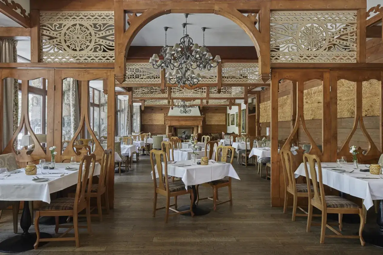 Elegancka sala restauracyjna z drewnianymi zdobieniami, idealna dla hoteli 5 gwiazdkowych. Stoły nakryte białymi obrusami, stylowe krzesła i ozdobny żyrandol tworzą luksusową atmosferę.