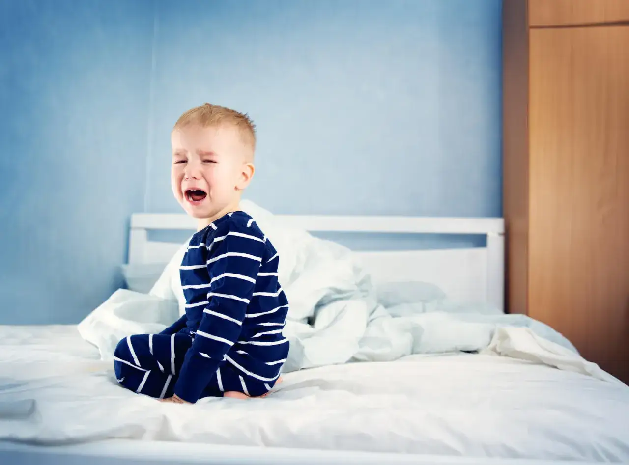 Bambino in pigiama a righe piange sul letto, svegliato da un pavor notturno.