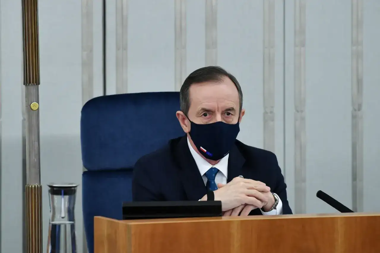 Tomasz Grodzki w maseczce z polską flagą, siedzi przy stole. Może właśnie odbierał list z Poczty Polskiej.