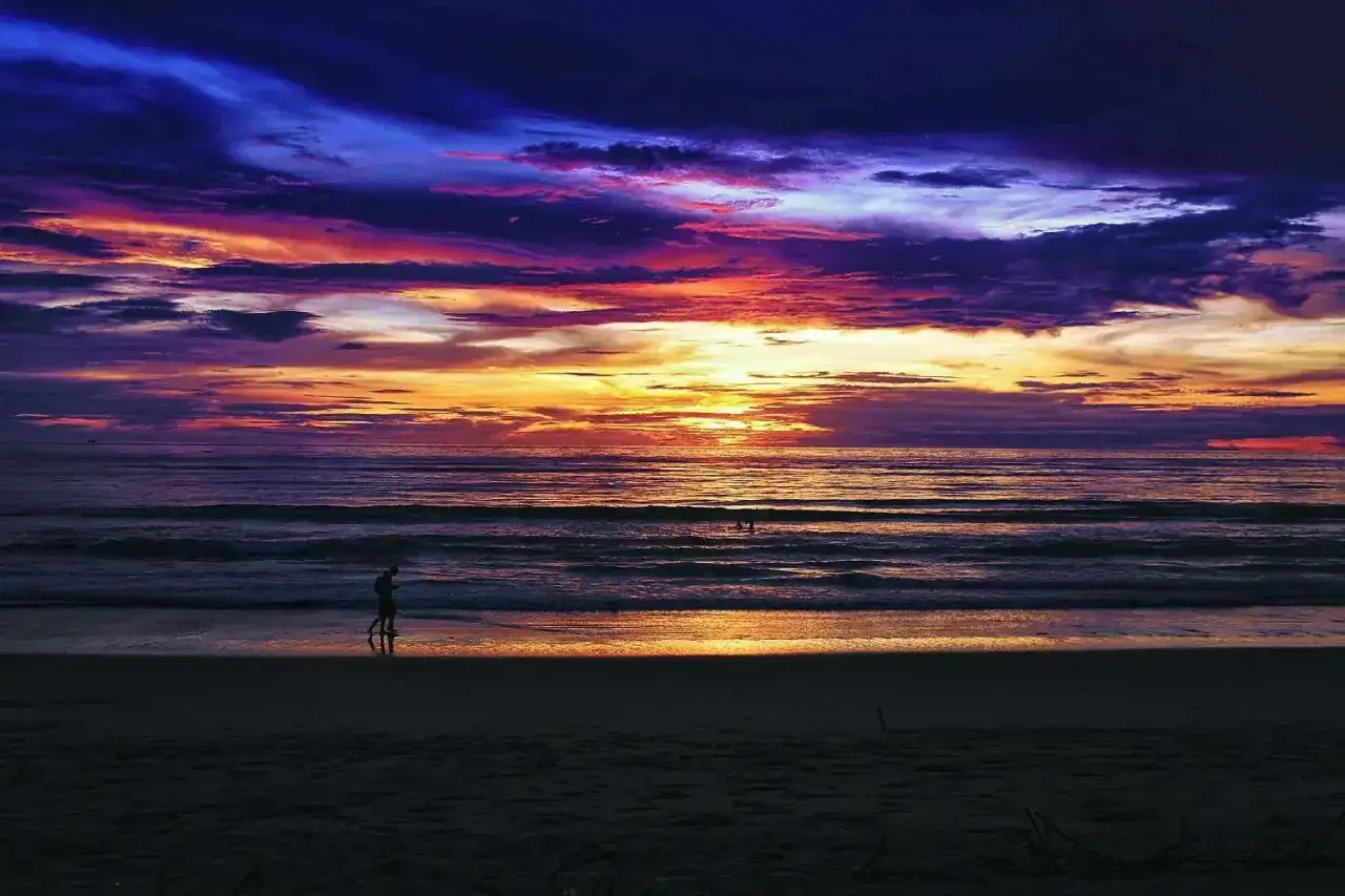 Ensam person promenerar p&aring; stranden vid en dramatisk solnedg&aring;ng i Thailand, Khao Lak. Himlen exploderar i lila och orange.