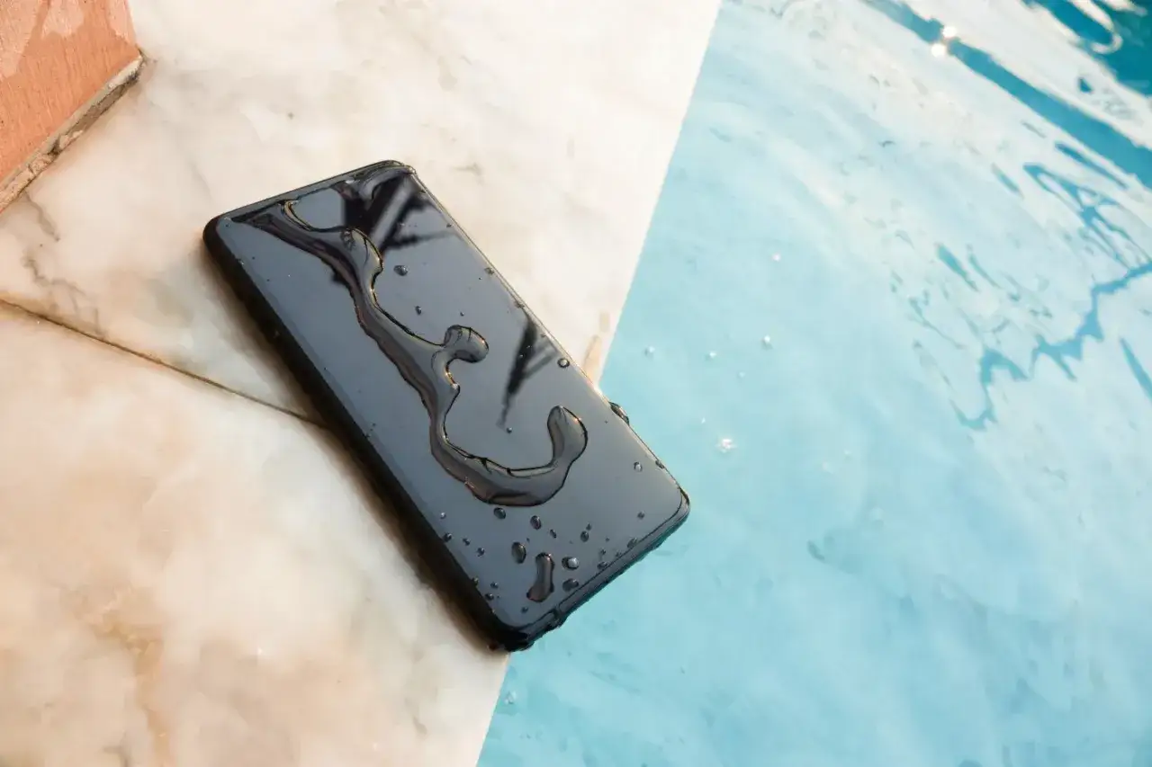 Czarny telefon leży na brzegu basenu, mokry od wody. Mimo że telefon wpadł do wody, nadal działa, co jest dowodem jego wodoodporności.