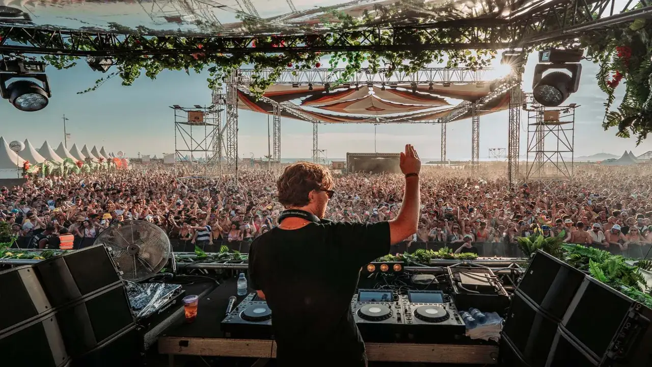 Un DJ, parmi les plus connus, salue une foule immense lors d'un festival en plein air. La sc&egrave;ne est d&eacute;cor&eacute;e de verdure, le soleil brille.