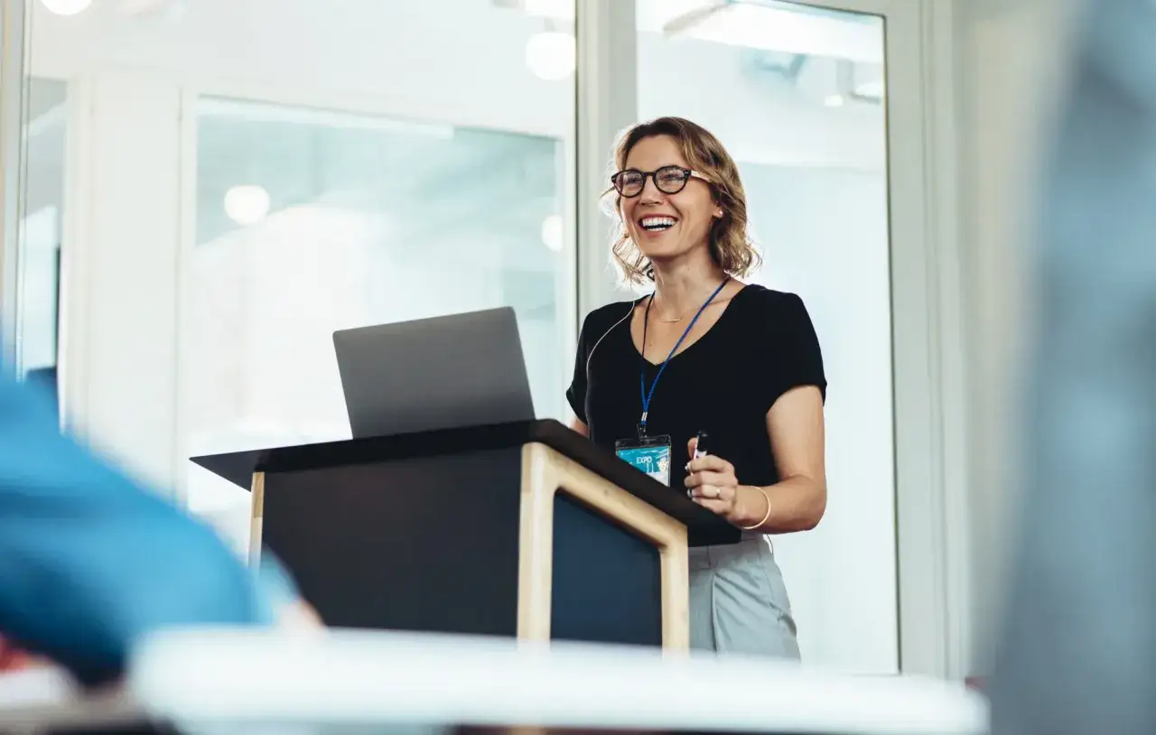 Een lachende vrouw geeft een presentatie. Ze staat achter een spreekgestoelte met een laptop. Waarom staren mensen naar een spreker? Misschien door haar enthousiasme.