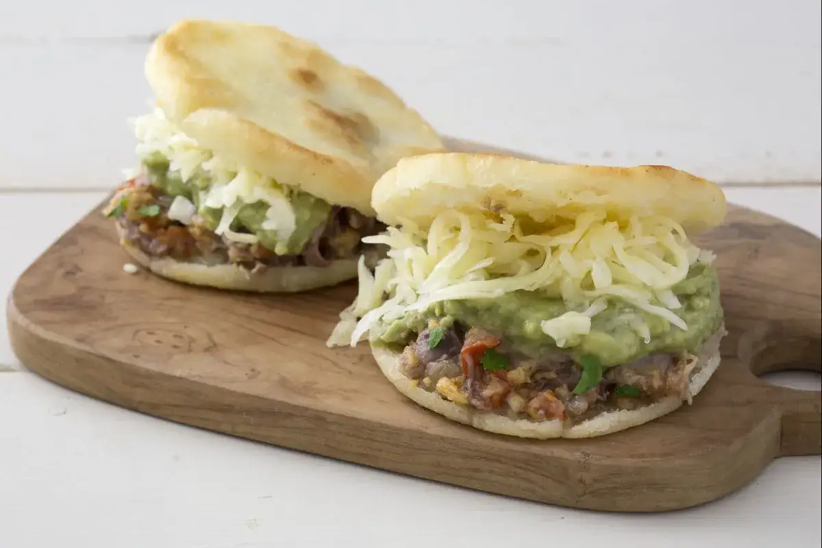Deux arepas garnies de guacamole, de viande effiloch&eacute;e et de fromage r&acirc;p&eacute;, servies sur une planche en bois.