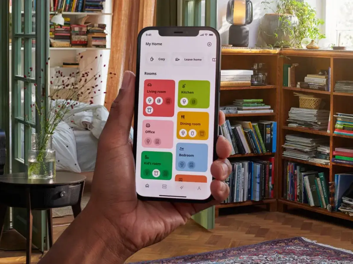 En hand h&aring;ller en mobil med en app f&ouml;r att dirigera hubben i hemmet. Appen visar rum som k&ouml;k och sovrum.