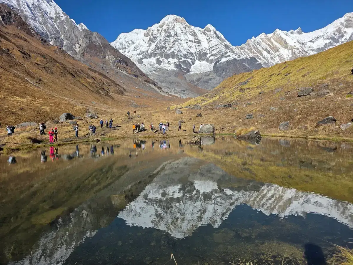 Grupa turystów podziwia odbicie ośnieżonych szczytów w spokojnej tafli jeziora. To widok z **Annapurna Base Camp**.