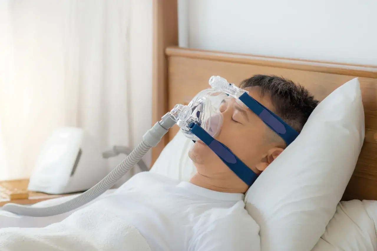 Mężczyzna śpi z maską CPAP, kt&oacute;ra pomaga przy chrapaniu. Warto skonsultować się z lekarzem w tej sprawie.