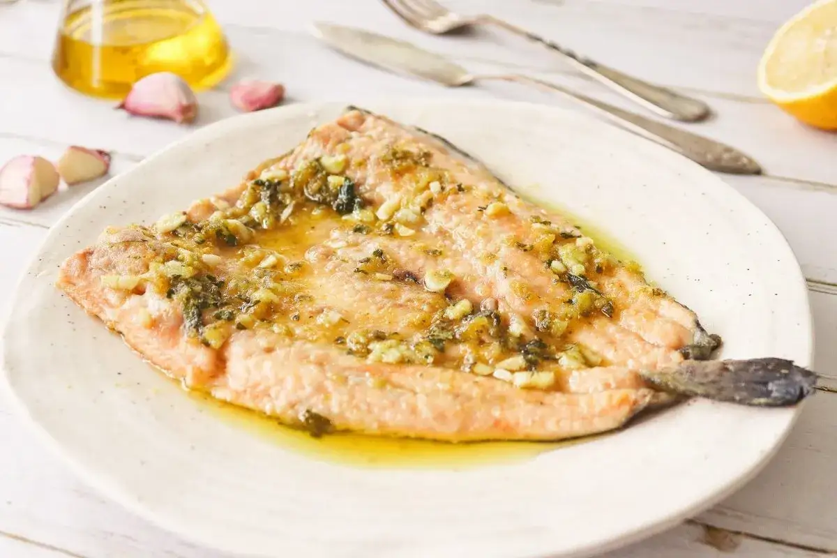 Trucha al ajillo servida en plato blanco, con aceite de oliva, ajos y limón de fondo.