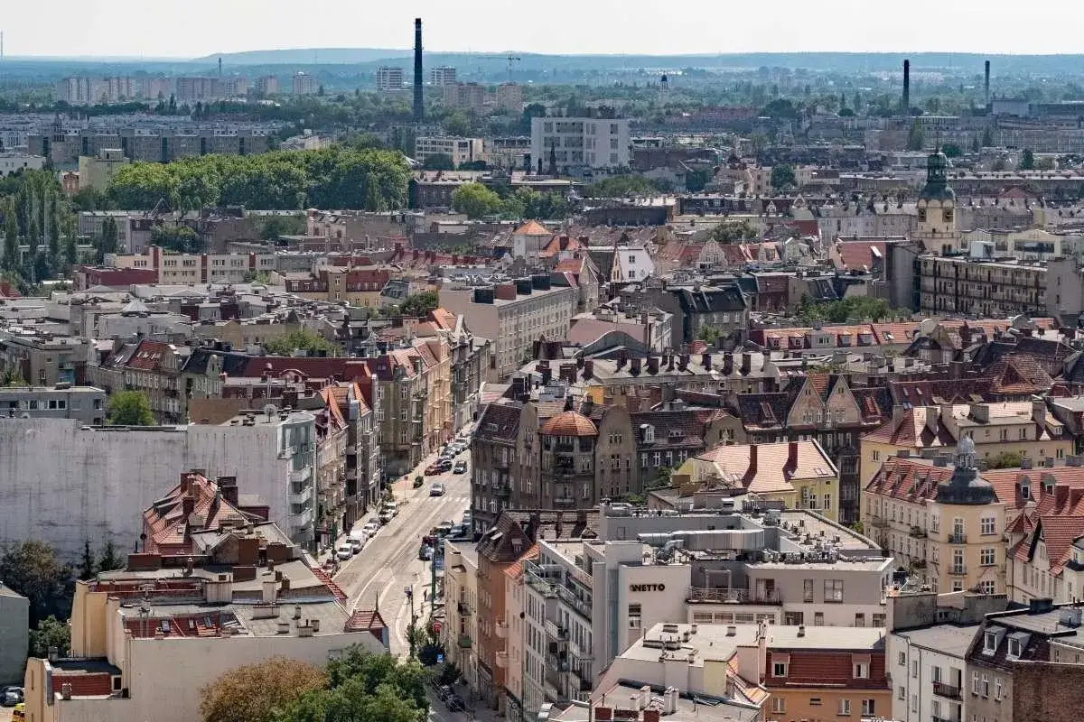 Widok na miasto z gęstą zabudową kamienic i dachów. W oddali widać kominy i zieleń, tworząc krajobraz wzgórza przemysła.