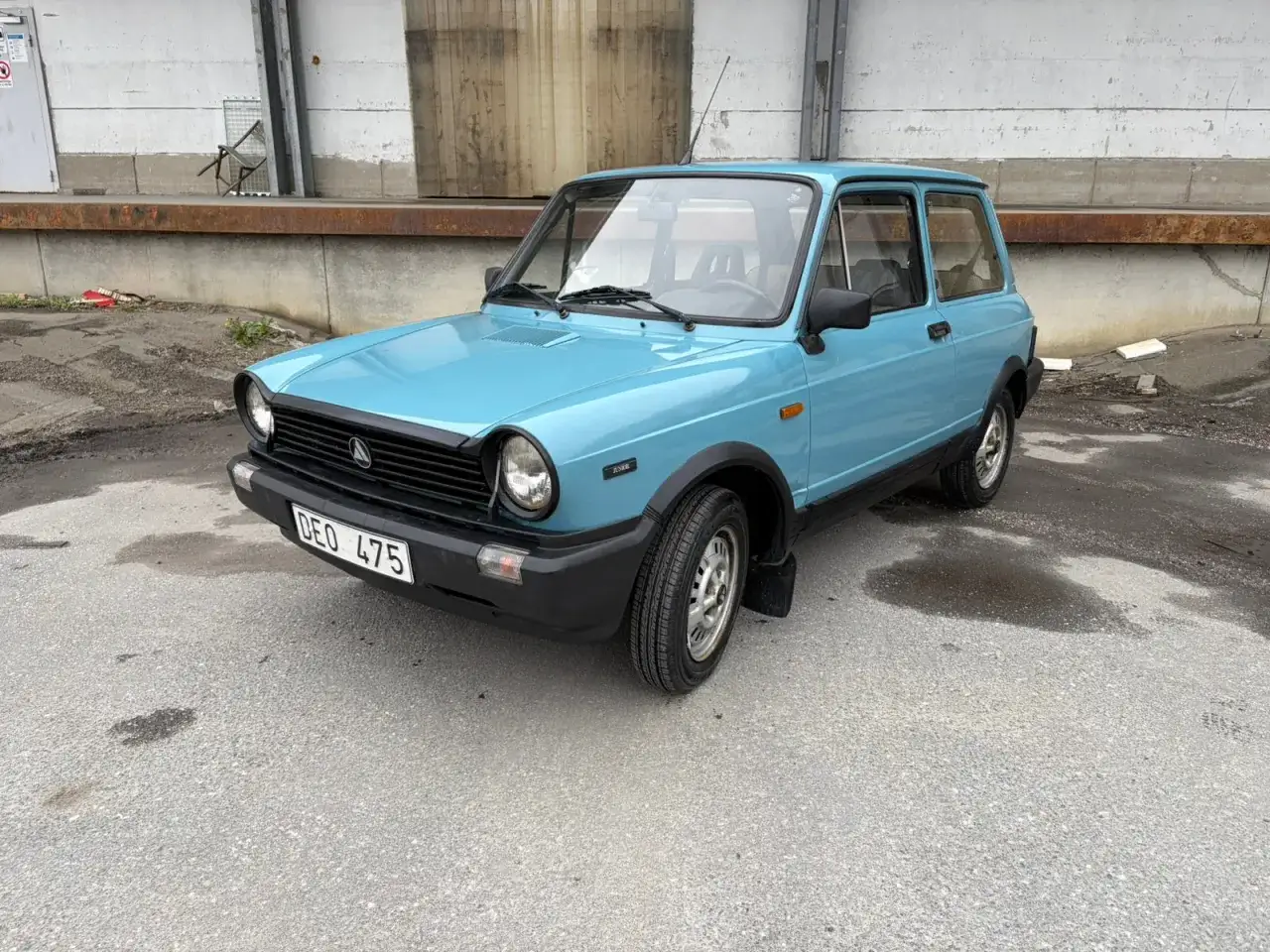 En ljusbl&aring; Lancia A112 Junior st&aring;r parkerad p&aring; en grusig yta framf&ouml;r en industribyggnad.