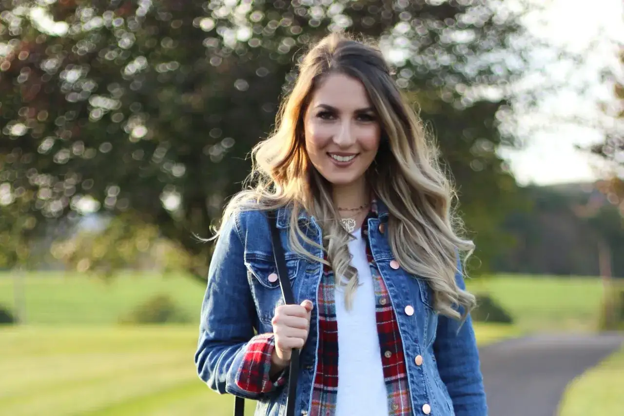 Frau mit blonden Haaren tr&auml;gt eine Jeansjacke, die sie l&auml;ssig mit einem karierten Hemd und einem wei&szlig;en T-Shirt kombiniert.