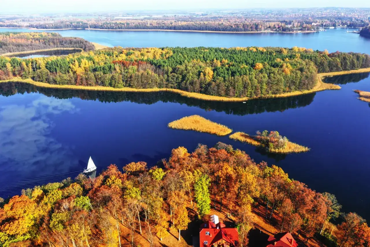 Jesienne barwy las&oacute;w nad jeziorem, idealne miejsce, gdzie na weekend listopadowy. Żagl&oacute;wka sunie po spokojnej tafli wody.