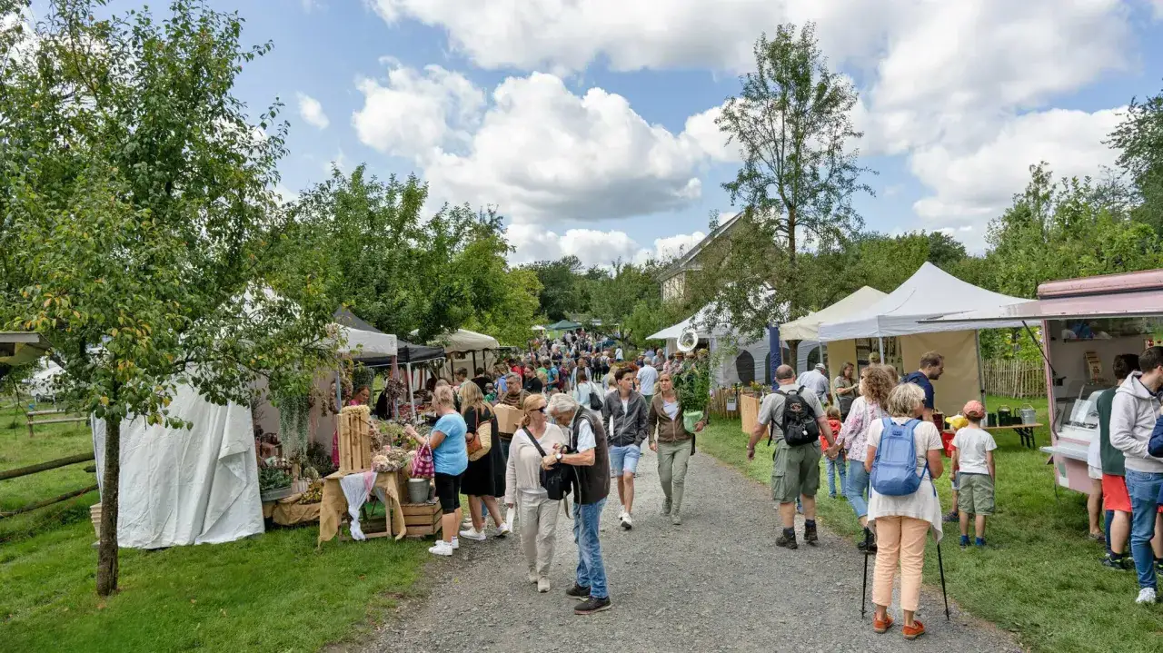 Menschen bummeln über den **Bauernmarkt Lindlar**, vorbei an Ständen mit Blumen und Lebensmitteln unter blauem Himmel.