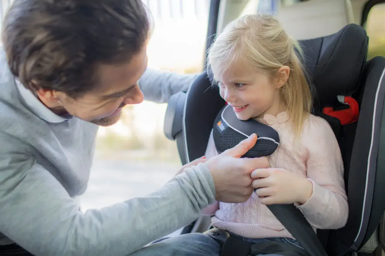 Padre ajusta el arnés de seguridad a su hija en el coche. Ella sonríe, lista para el viaje. Pregunta: ¿cuándo puede ir un niño sin silla en el coche?
