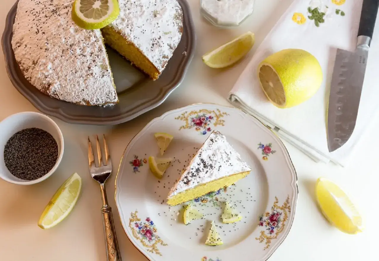 Una fetta di torta al limone, spolverata di zucchero a velo e semi di papavero, su un piatto decorato. Limoni freschi e un coltello completano la scena.
